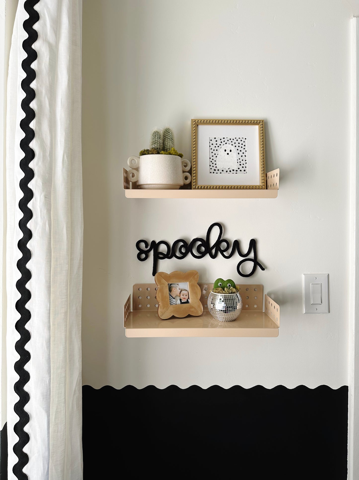 Black knitted wire ‘Spooky’ sign between two wooden shelves in a boy’s bedroom, styled with small plants and picture frames, white curtains with black rickrack, and a black-and-white wavy wall border for a playful Halloween vibe.