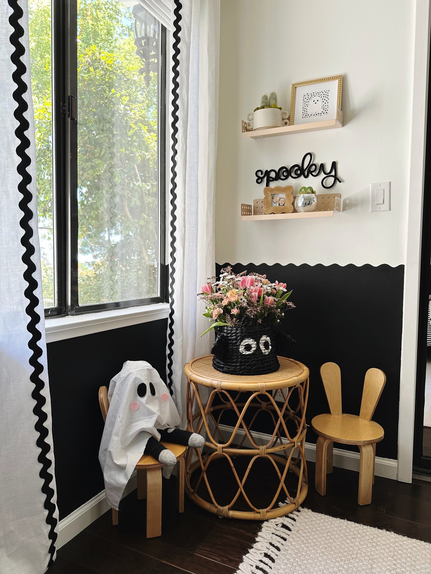 Black knitted wire ‘Spooky’ sign hanging between two wooden shelves above a children’s table with light wood chairs featuring bunny ears in a boy’s bedroom. Room has white curtains with black rickrack, a black-and-white wavy wall border, and shelves styled with small plants and picture frames, creating a playful, modern Scandinavian Halloween vibe.
