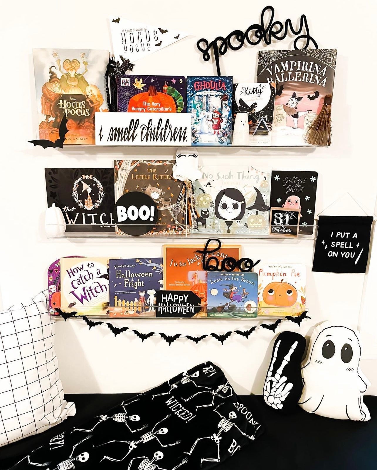 Children’s bedroom wall with three layered bookshelves filled with Halloween-themed books featuring pumpkins, witches, vampires, and ghosts. Black knitted wire word signs spelling ‘Spooky’ and ‘Boo’ are displayed on the shelves. Ghost garlands are draped across the shelves, and Halloween-themed accent pillows create a cozy reading spot with cushions below. Playful, festive, and whimsical seasonal décor for a child’s room or nursery.