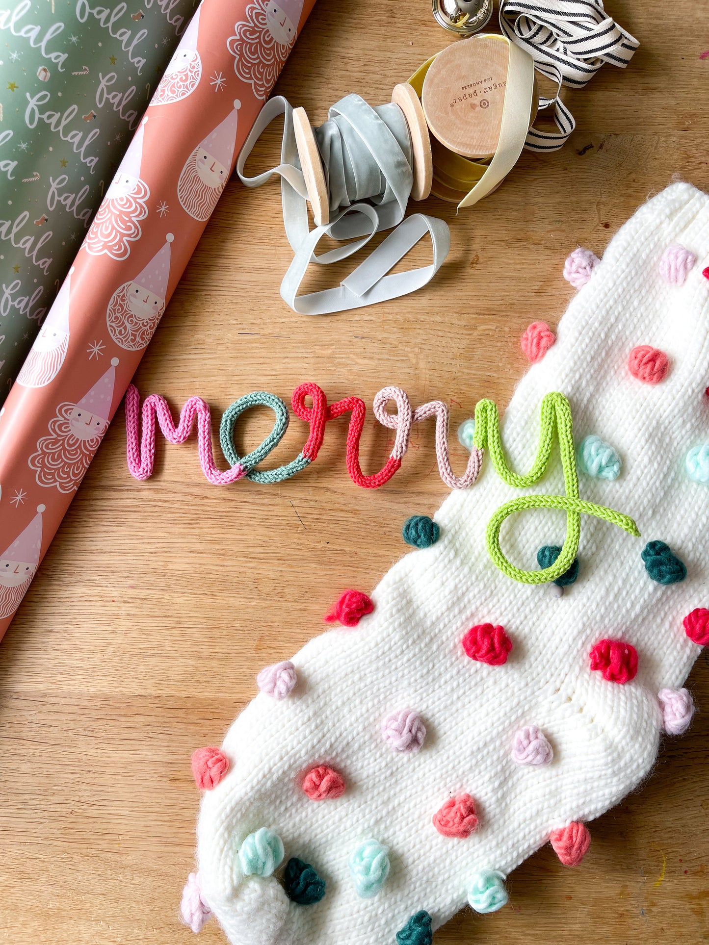 A handmade 'Merry' sign with multicolored buttons and ribbons on a wooden surface, with Christmas-themed decorations in the background.