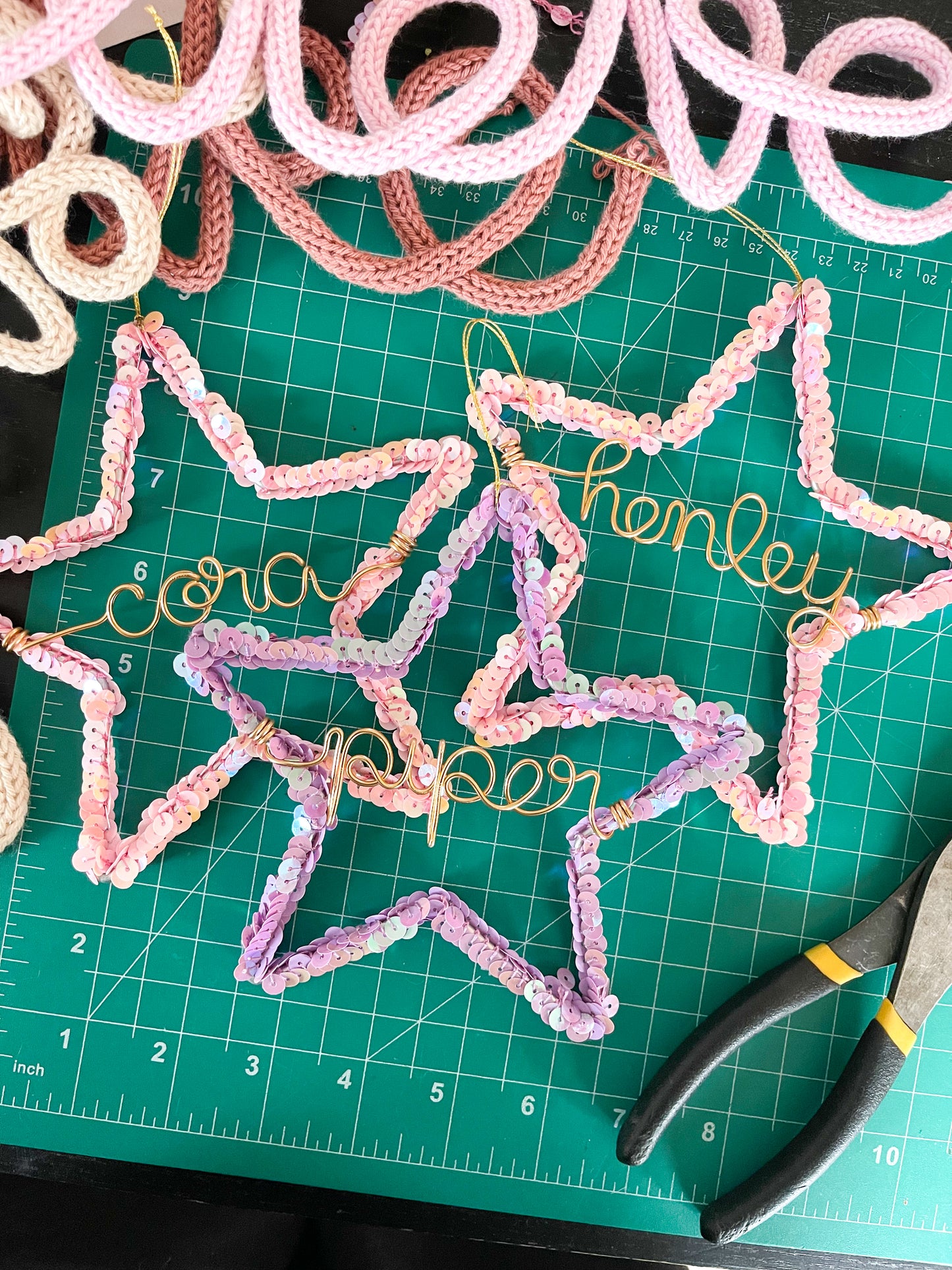 Three handmade sequin star ornaments on a green cutting mat — pink stars personalized for Cora and Henley, and a purple star for Piper — ready to be packaged as Christmas gifts, with bits of other wire names peeking into the frame.