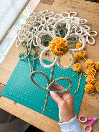 A handmade rope flower decoration with a yellow pom-pom center and green rope stem, held by a hand over a teal cutting mat on a wooden table with other rope flowers and yarn.
