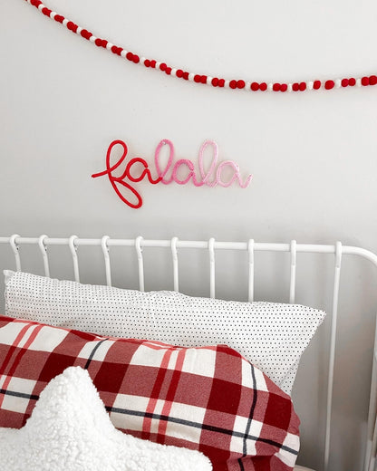 Red to  pink ombre knitted wire name sign 'fa la la' in color block style. Hanging on wall above child's bed with plaid holiday sheets.