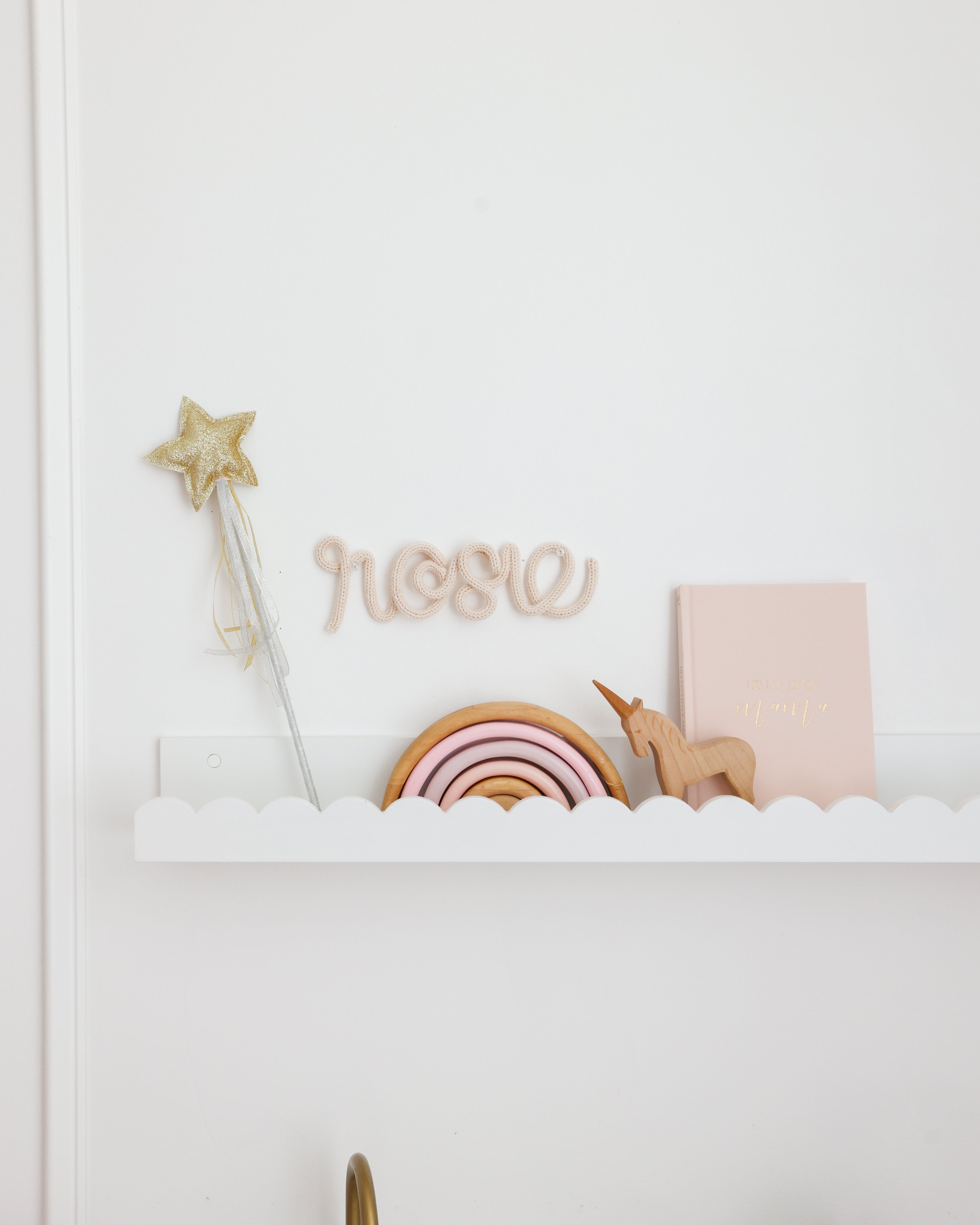 A styled nursery shelf display featuring a wooden name sign spelling 'rose', a glittery gold star wand, a wooden rainbow, a small wooden unicorn, and a blush pink hardcover book on a white shelf against a white wall.