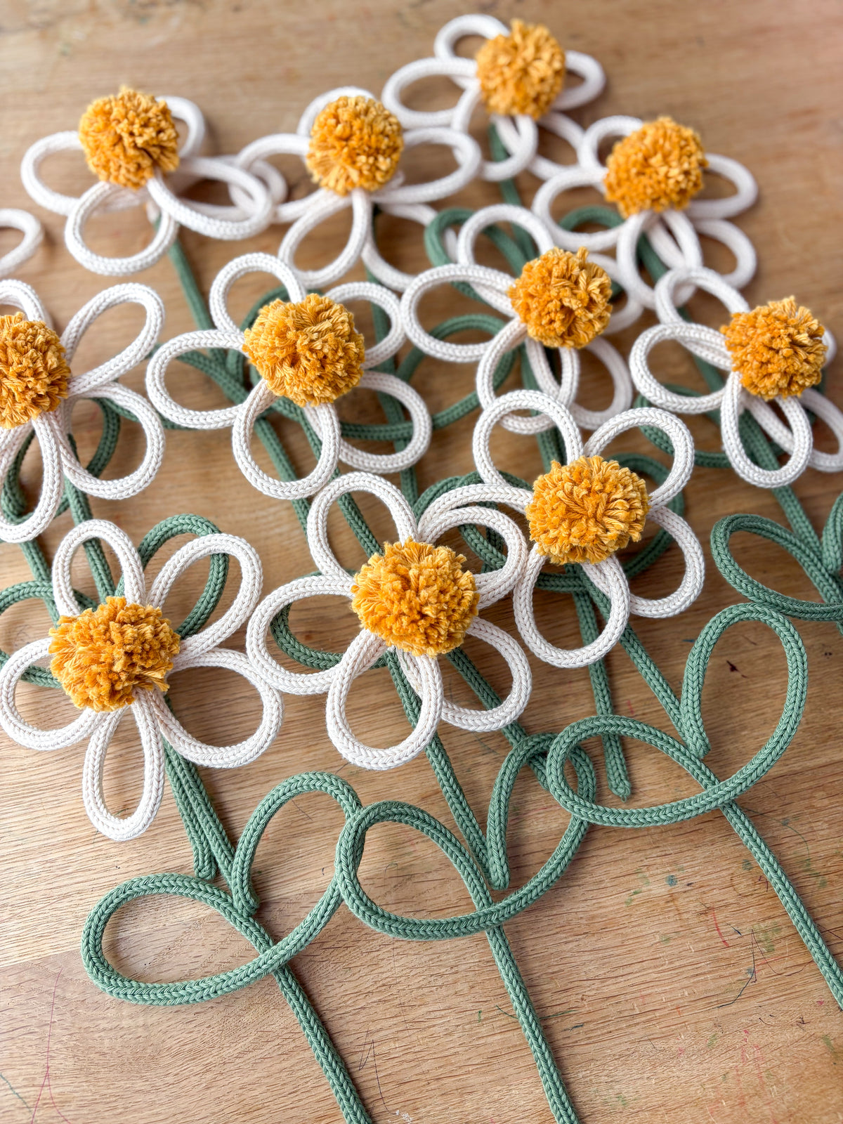 A close-up of several decorative rope flowers laid on a wooden surface. Each flower has knitted rope/yarn petals and a textured mustard-yellow center, with green rope stems extending downward.