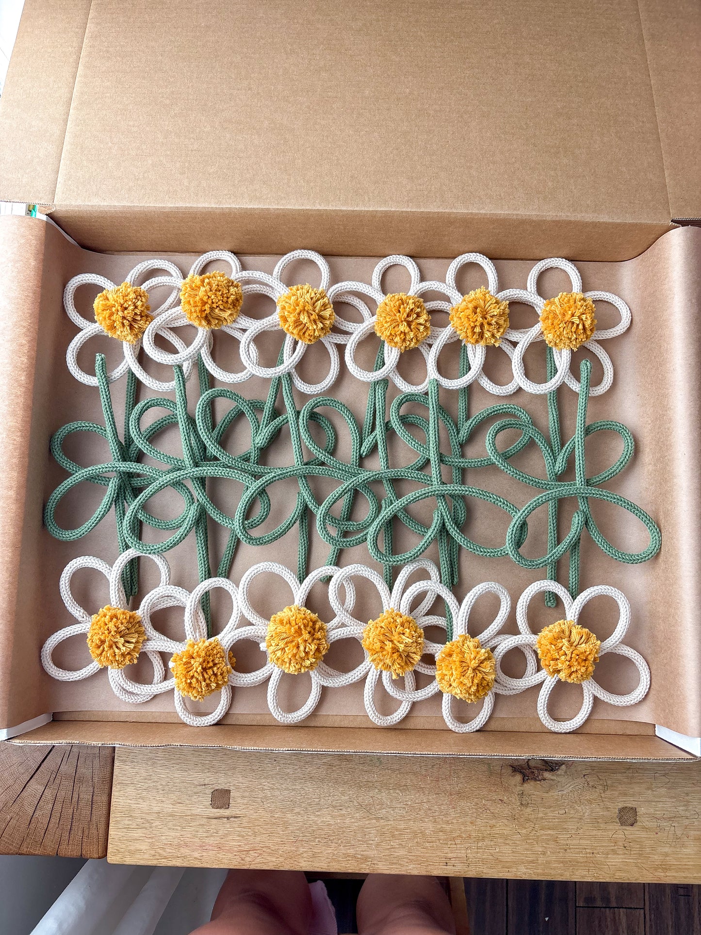 A set of handmade wire flower s with white petals, yellow pom-pom centers, and green stems, displayed inside an open brown cardboard box.