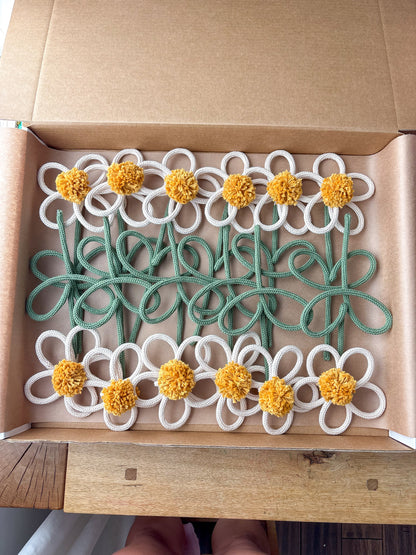 A set of handmade wire flower s with white petals, yellow pom-pom centers, and green stems, displayed inside an open brown cardboard box.