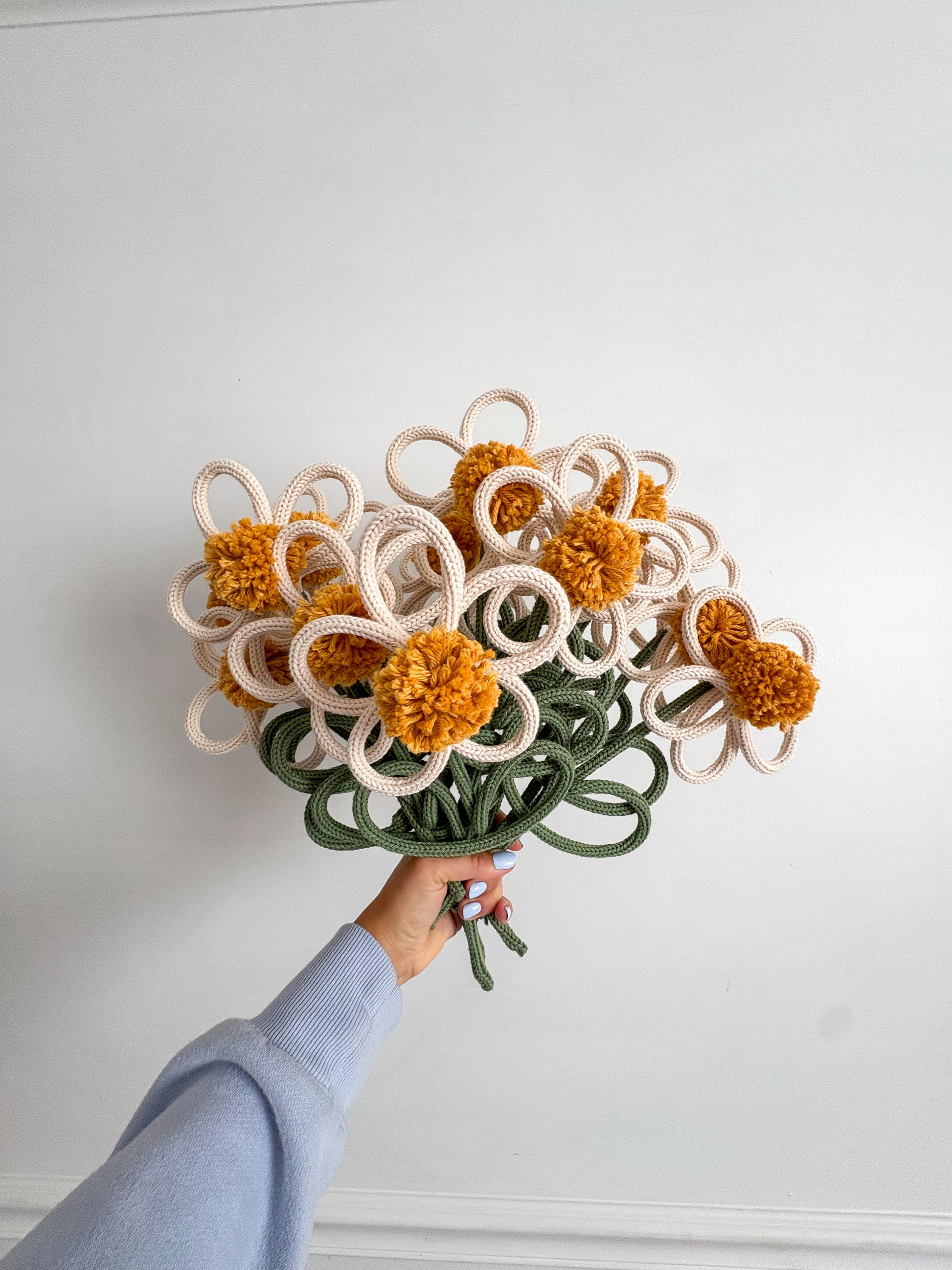 A hand holding a bouquet of handmade knitted flowers with yellow pom-pom centers, cream petals, and green stems against a plain white wall.
