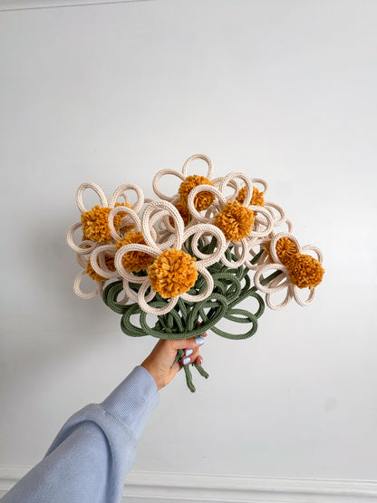 A hand holding a bouquet of handmade knitted flowers with yellow pom-pom centers, cream petals, and green stems against a plain white wall.