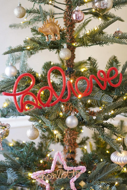 A decorated Christmas tree with red yarn knitted wire word that spell out the word 'Believe' and a pink star ornament with the word 'Charlotte' written on it.