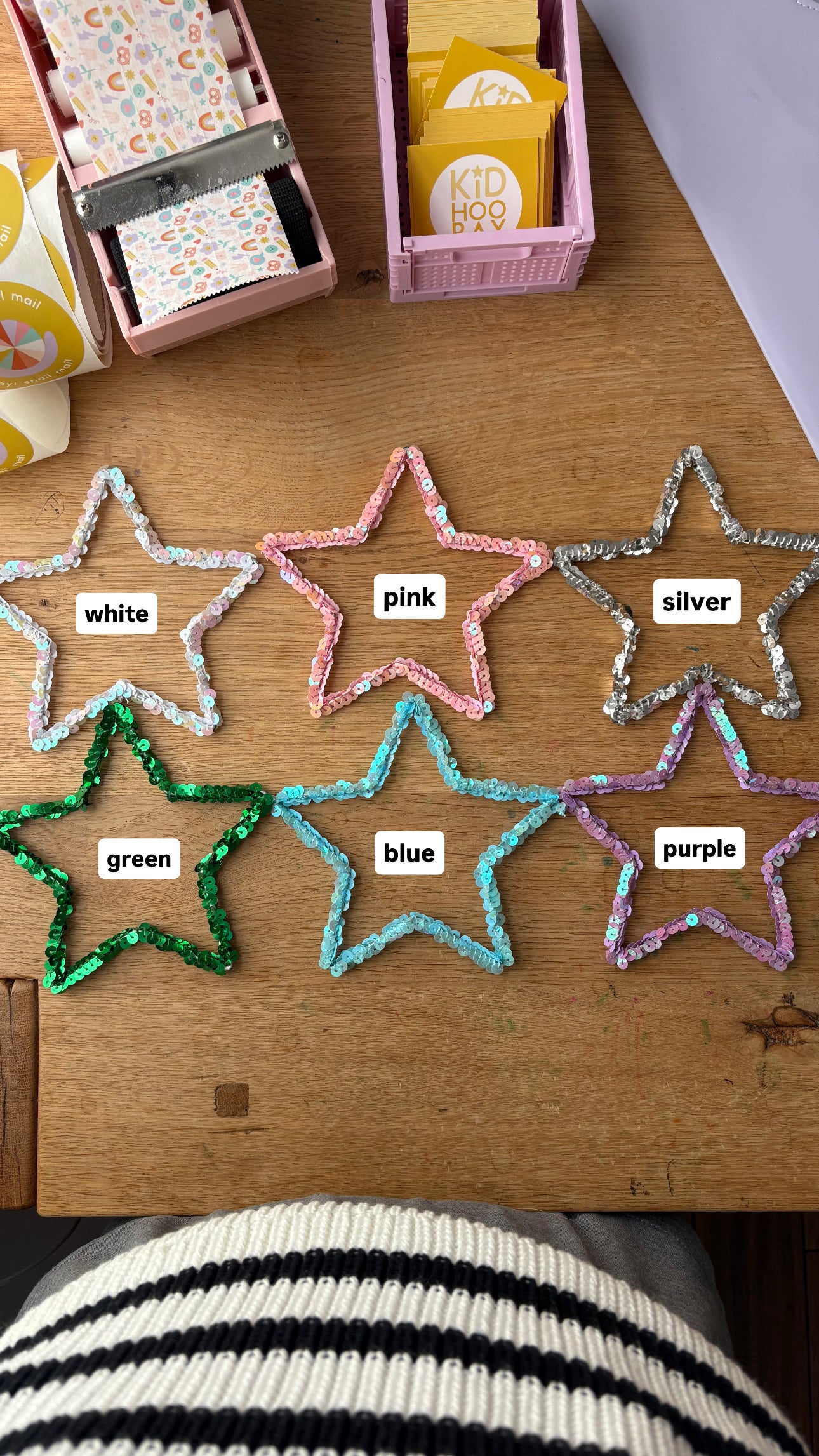 Six sequin star ornaments on a wooden table showing color options with labels — white, pink, purple, blue, silver, and green — handmade personalized Christmas ornaments color chart.