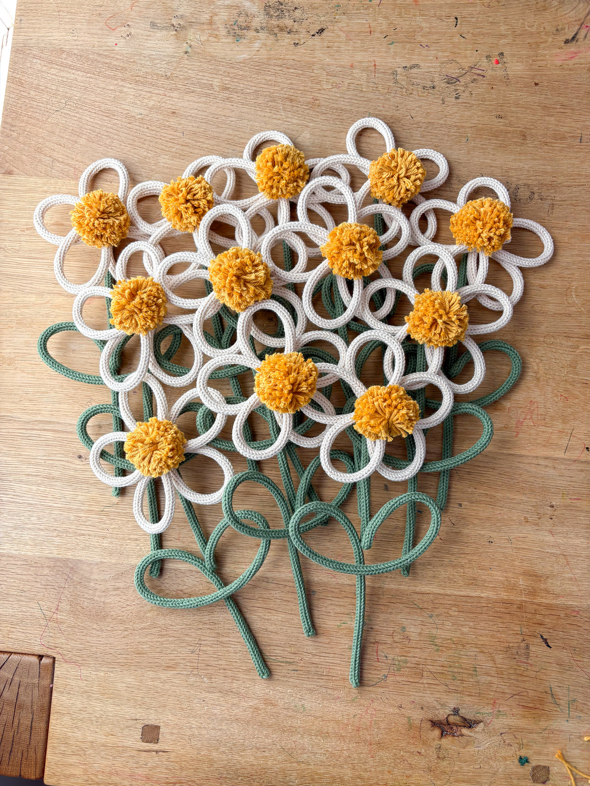 A cluster of decorative rope flowers arranged on a wooden tabletop. Each flower has a white rope petal, a yellow pom-pom center, and a green rope stem.