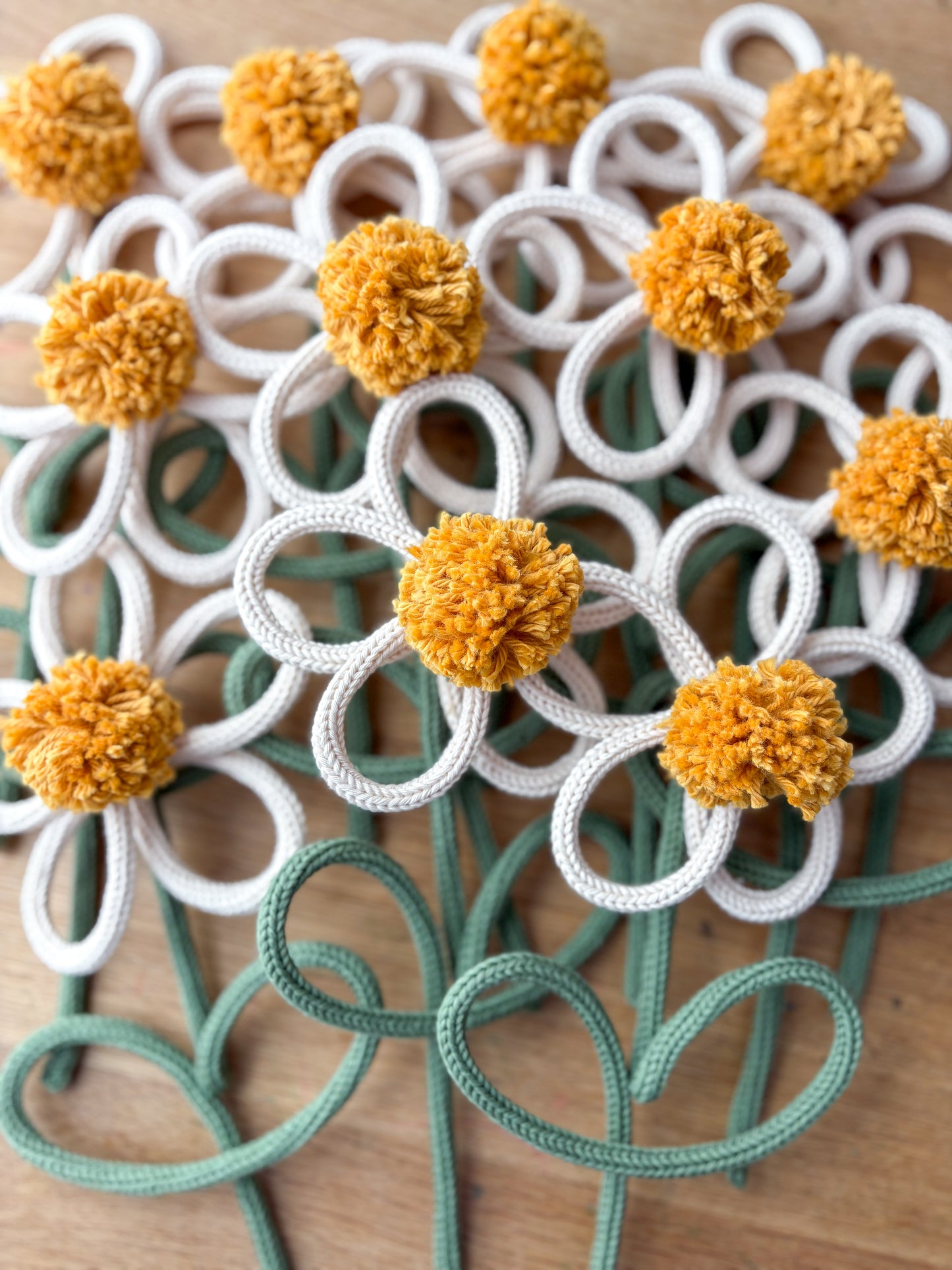A close-up of handmade rope/yarn flower with white petals, mustard yellow pom-pom centers, and sage green stems on a wooden surface.