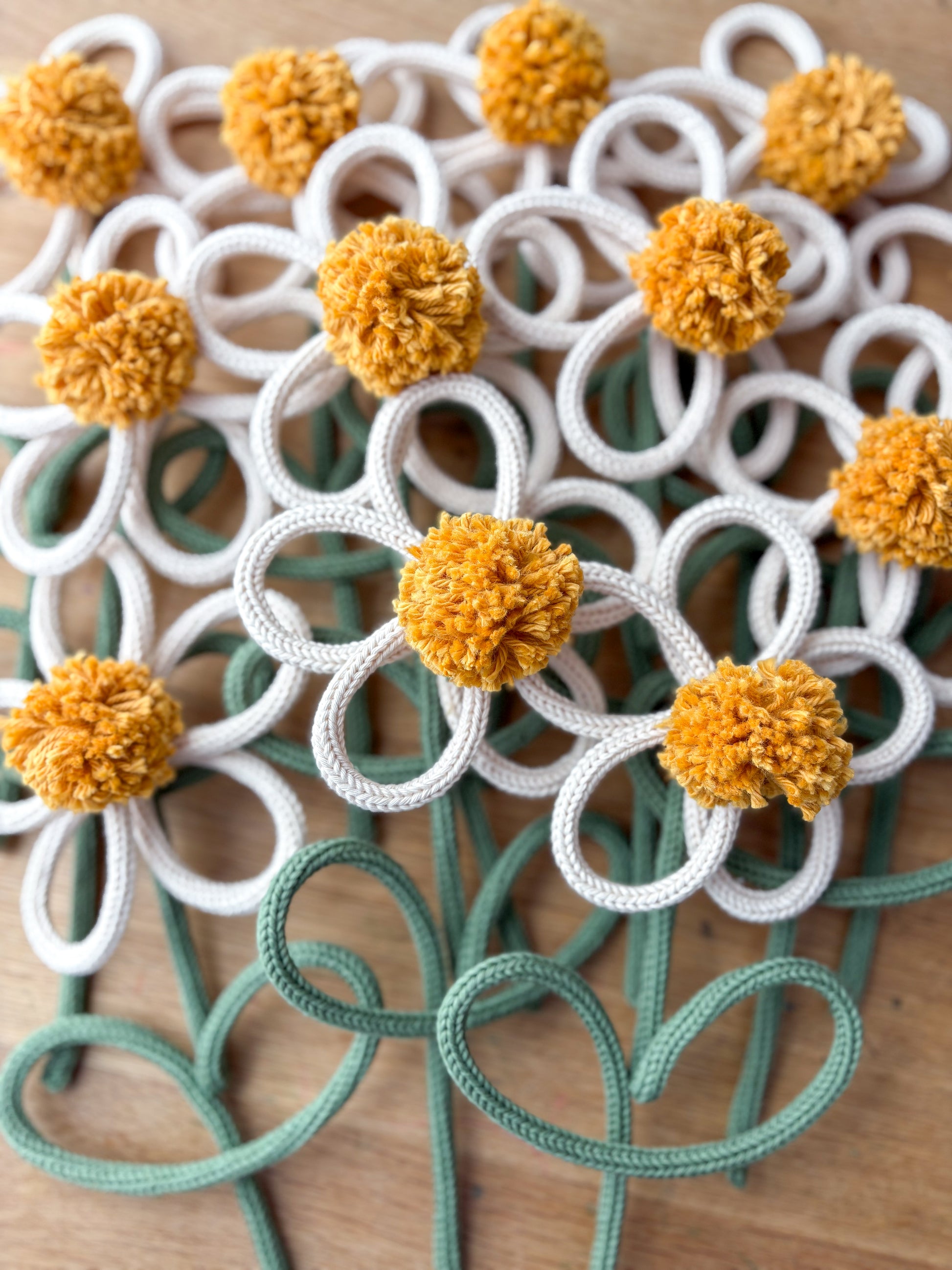A close-up of handmade rope/yarn flower with white petals, mustard yellow pom-pom centers, and sage green stems on a wooden surface.