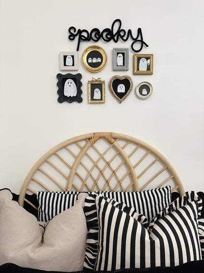 Black knitted wire ‘Spooky’ sign above a black-and-white ghost gallery wall in a boy’s bedroom with striped pillows and a woven headboard, creating a playful Halloween vibe.