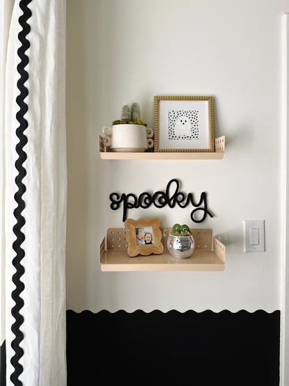 Black knitted wire ‘Spooky’ sign between two wooden shelves in a boy’s bedroom, styled with small plants and picture frames, white curtains with black rickrack, and a black-and-white wavy wall border for a playful Halloween vibe.