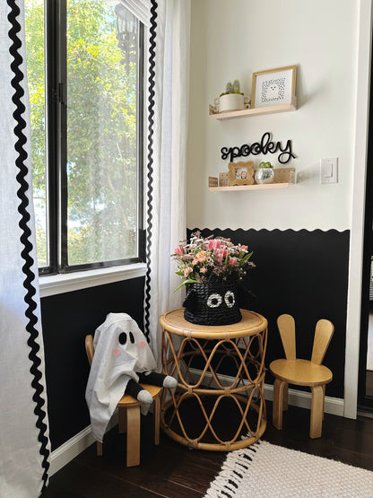 Black knitted wire ‘Spooky’ sign hanging between two wooden shelves above a children’s table with light wood chairs featuring bunny ears in a boy’s bedroom. Room has white curtains with black rickrack, a black-and-white wavy wall border, and shelves styled with small plants and picture frames, creating a playful, modern Scandinavian Halloween vibe.