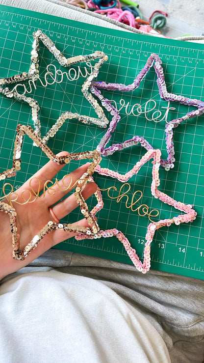 Hand holding a bronze sequin star ornament personalized with the name Kelsie, with three other wire star ornaments on a green cutting mat — silver for Andrew, pink for Chloe, and purple for Violet - handmade Christmas decorations.