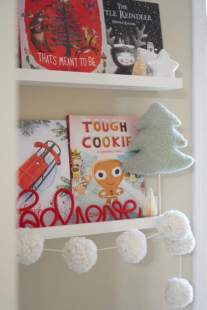 White shelf with Christmas children’s books ‘The Little Reindeer,’ ‘That’s Meant to Be,’ and ‘Tough Cookie,’ a green plush Christmas tree, and a red rope sign spelling ‘believe.’