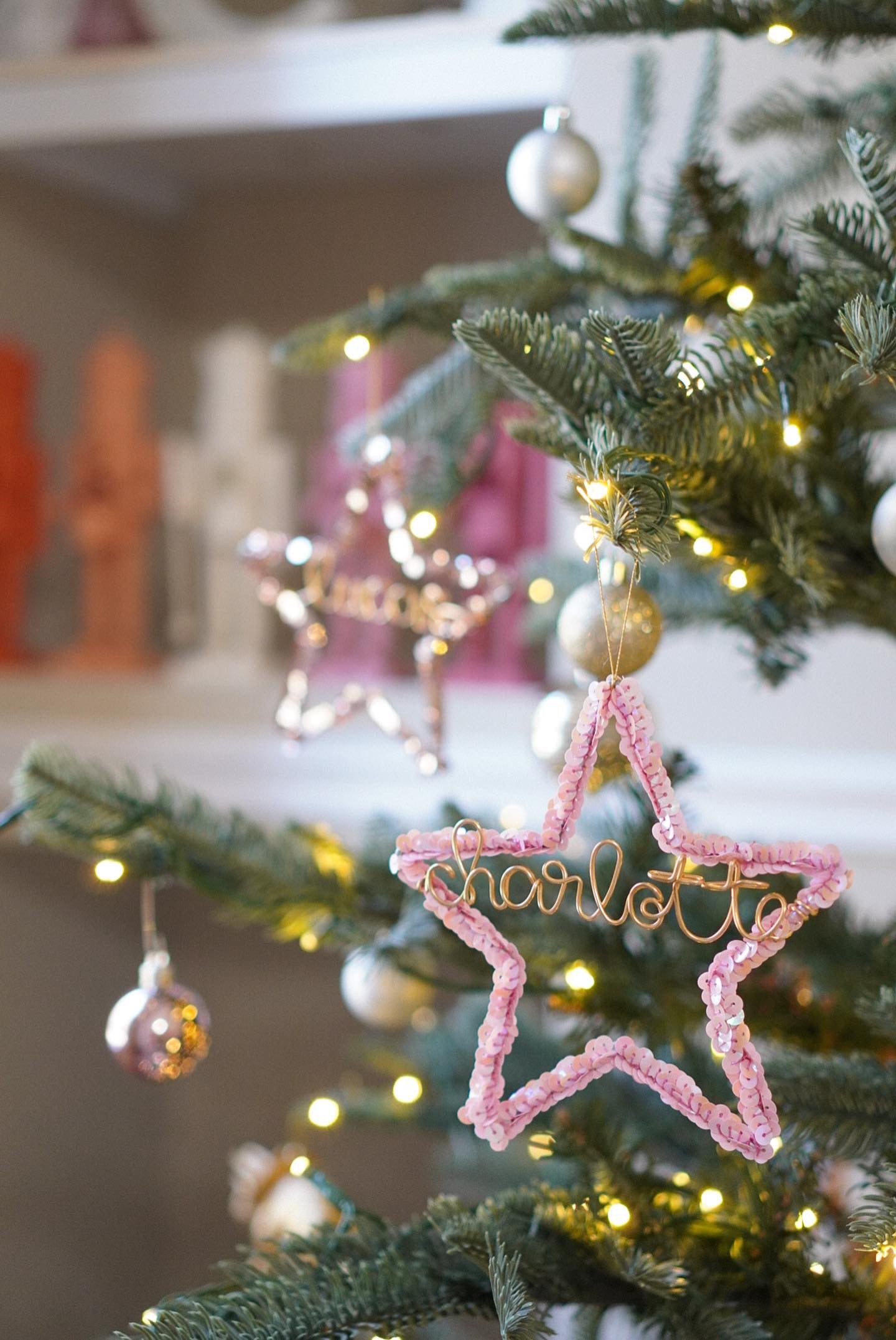Pink sequin star ornament personalized with the name Charlotte in gold wire, hanging on a Christmas tree with another Luxe Star in the background.