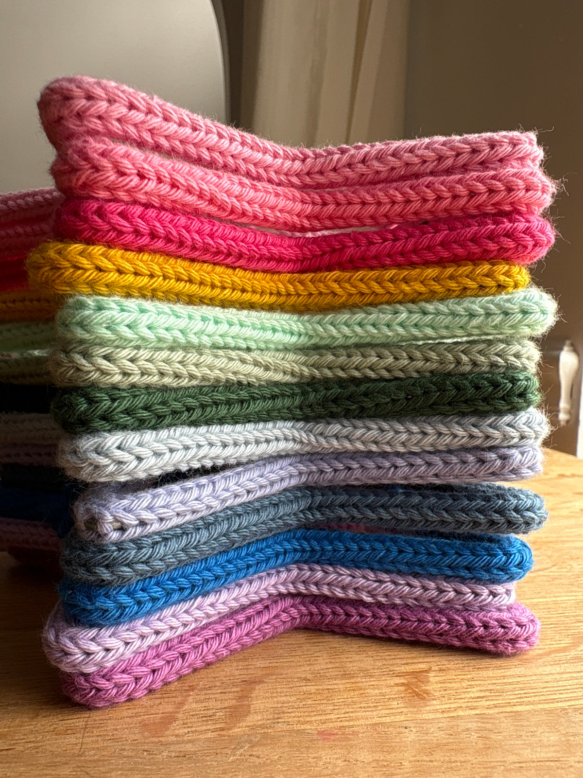 A stack of colorful crocheted or knit fabric pieces, likely dishcloths or coasters, arranged in a curved stack on a wooden table. The top piece is pink, with layers of bright pink, yellow, mint green, olive green, light gray, lavender, blue, and magenta below.