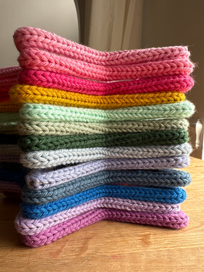 A stack of colorful crocheted or knit fabric pieces, likely dishcloths or coasters, arranged in a curved stack on a wooden table. The top piece is pink, with layers of bright pink, yellow, mint green, olive green, light gray, lavender, blue, and magenta below.
