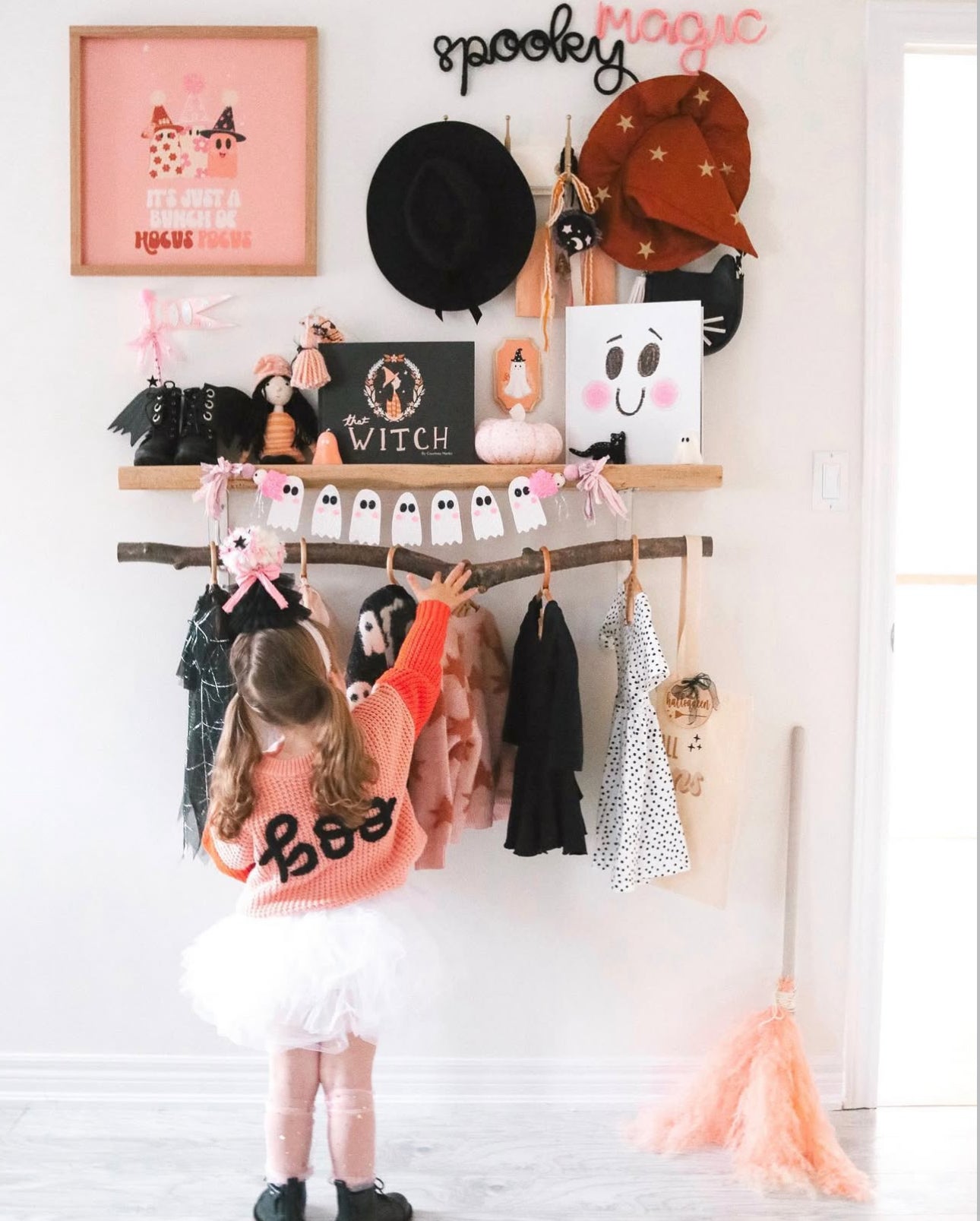 Little girl wearing a ‘Boo’ sweater with her back to the camera, reaching up to a shelf decorated with whimsical Halloween accents. Shelf features black knitted wire word signs spelling ‘Spooky’ and ‘Magic,’ along with Halloween-themed books about witches and pumpkins, a cat-shaped purse, and festive wall art. Below the shelf hangs a rod with Halloween-colored clothes and dress-up pieces. Light peach, pink, black, and white tones create a playful, girly, and seasonal vibe.