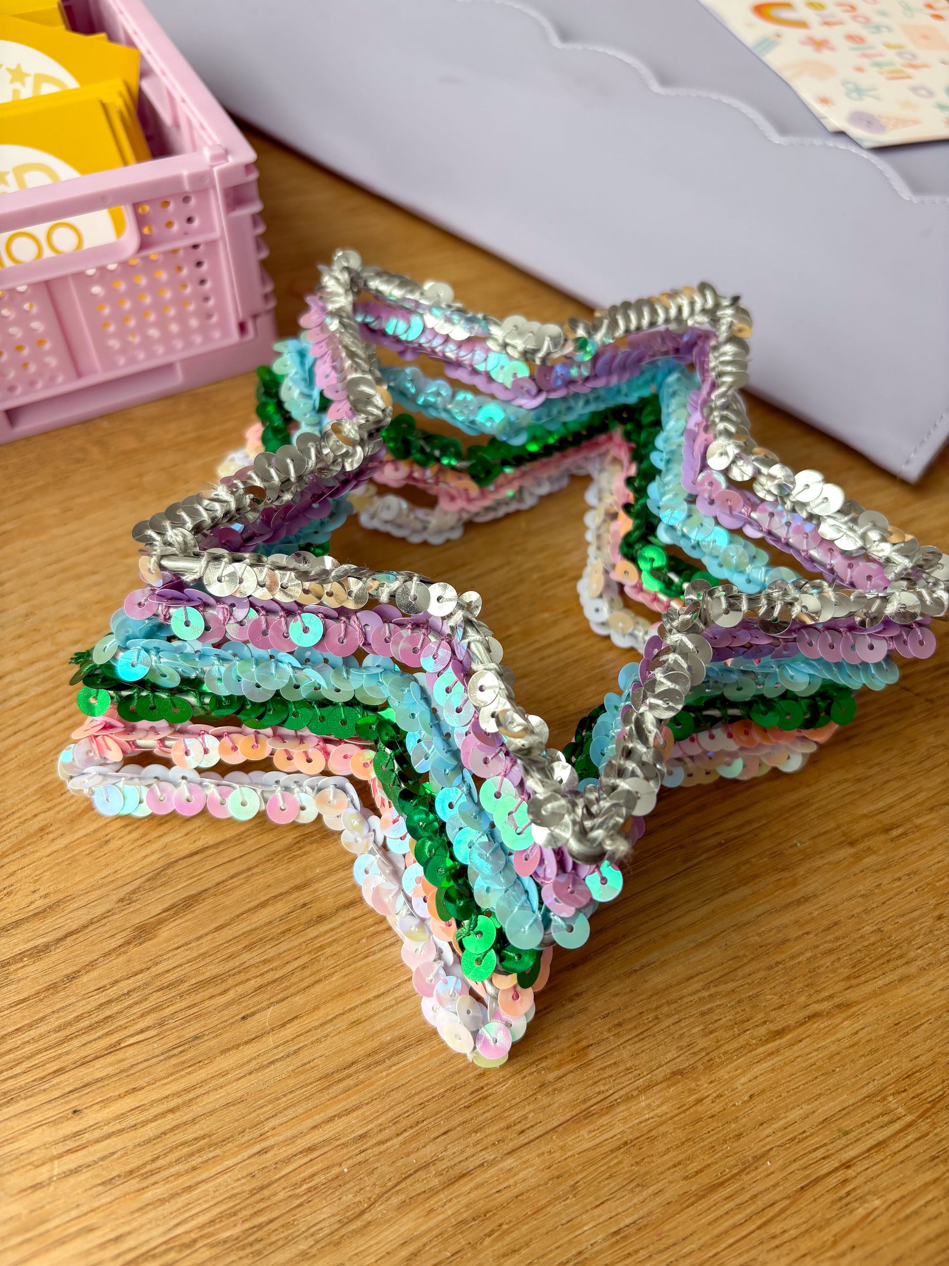 Stack of six sequin star ornaments in different colors — white, pink, purple, blue, silver, and green — displayed on a wooden surface, handmade personalized Christmas ornaments color samples.