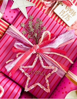Flatlay of pink-wrapped Christmas gifts adorned with embellishments and gift toppers, including a personalized pink sequin star ornament with gold wire name Cora, perfect for little girls’ holiday presents.