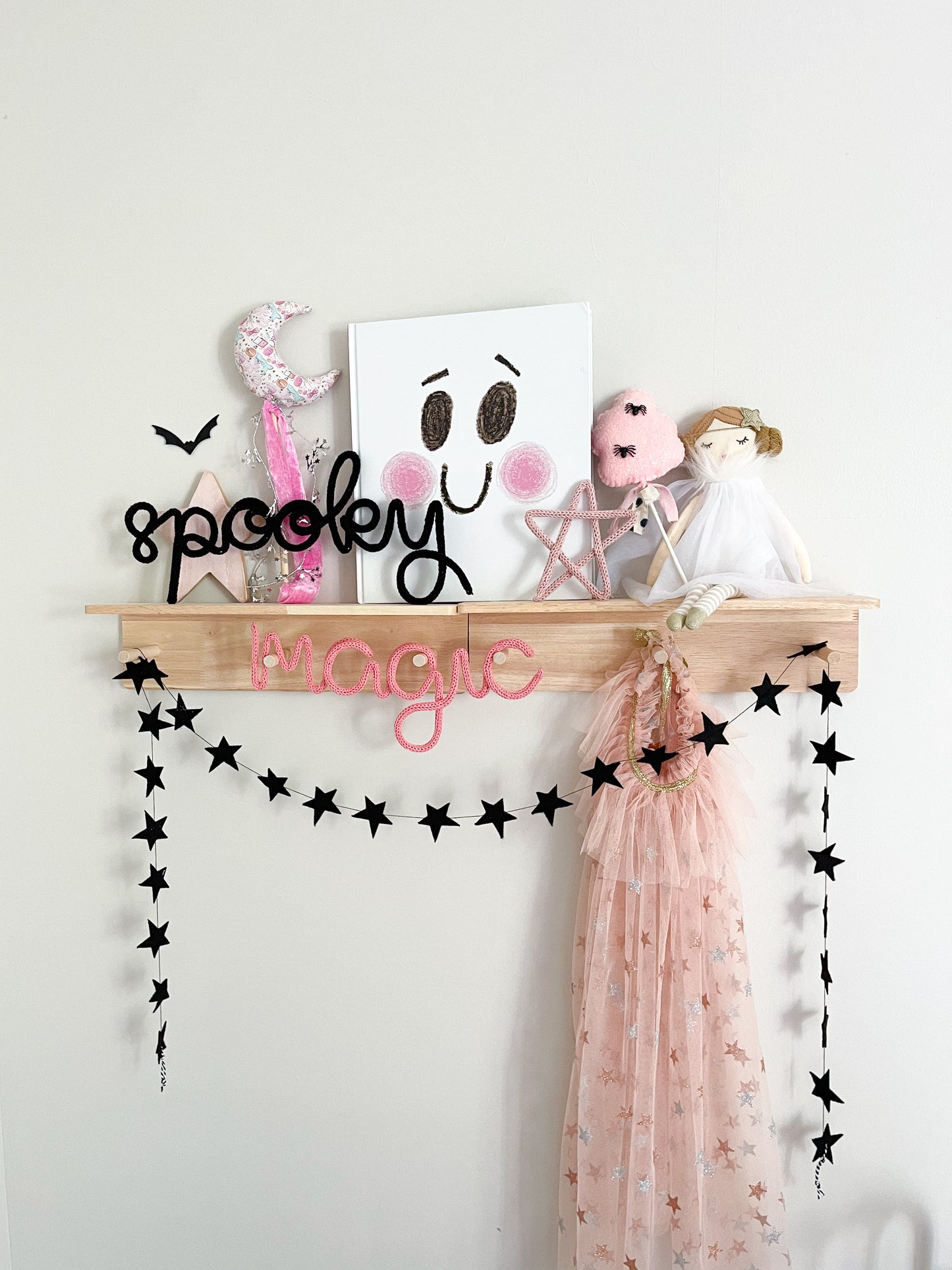 Whimsical Halloween décor styled on a peg rail shelf in a little girl’s bedroom, featuring black knitted wire word signs that spell ‘Spooky’ and ‘Magic.’ Shelf is decorated with playful star accents, a black star garland, a moon wand, a Scandinavian-style doll, and a cotton candy wand with tiny spiders. Pink, white, and black color scheme creates a soft yet festive vibe, with a children’s book featuring a ghost cover adding to the magical, seasonal look.