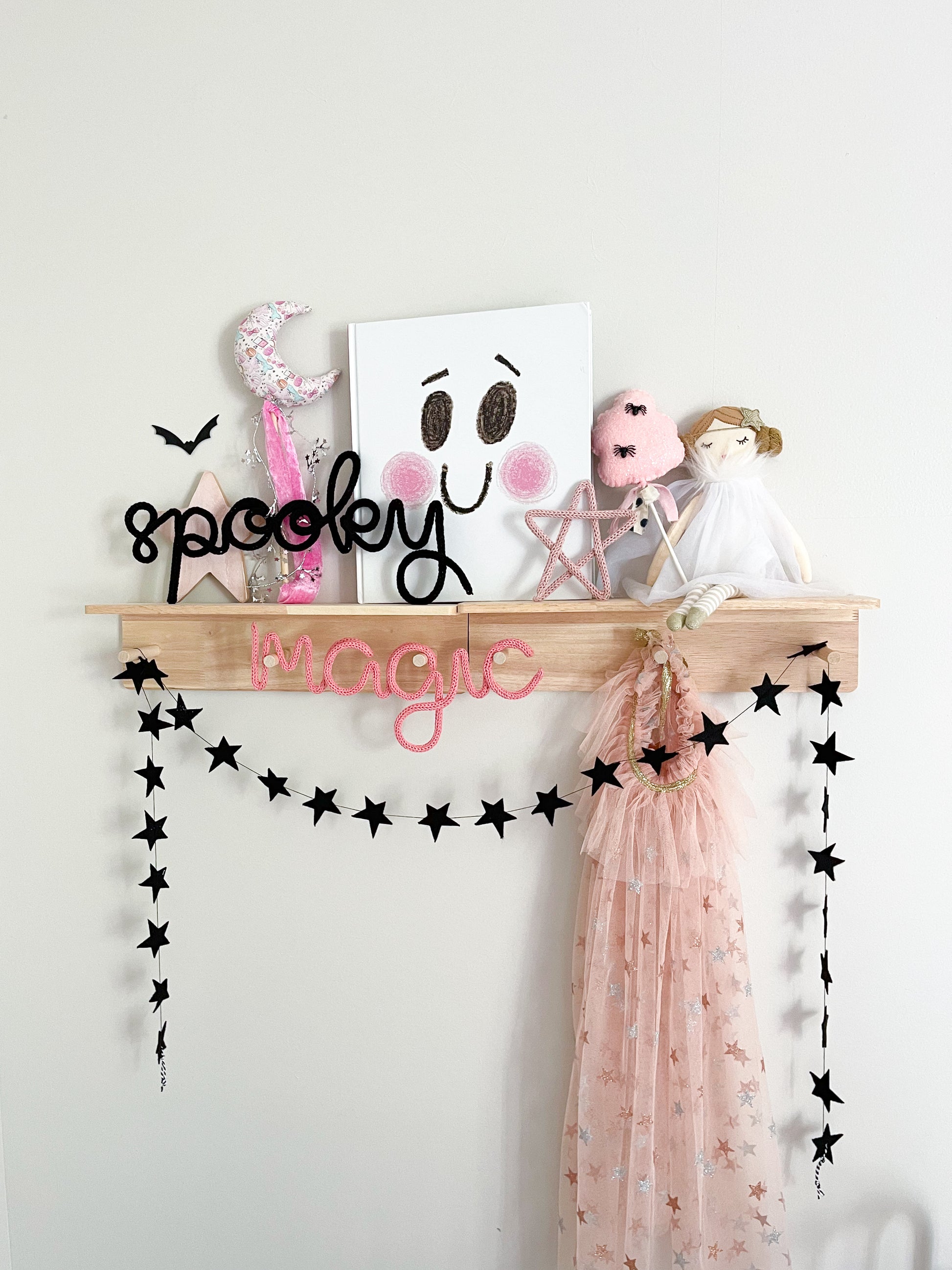Whimsical Halloween décor styled on a peg rail shelf in a little girl’s bedroom, featuring black knitted wire word signs that spell ‘Spooky’ and ‘Magic.’ Shelf is decorated with playful star accents, a black star garland, a moon wand, a Scandinavian-style doll, and a cotton candy wand with tiny spiders. Pink, white, and black color scheme creates a soft yet festive vibe, with a children’s book featuring a ghost cover adding to the magical, seasonal look.