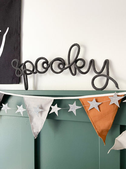Handmade black knitted wire word sign that spells ‘Spooky,’ propped on green board and batten wall in a boy’s bedroom. Behind the sign are layered decorations including a star garland and a neutral-colored bunting banner, creating a playful yet stylish Halloween setup. This kids’ room wall décor is festive but subtle, making it perfect for a seasonal touch in a nursery, playroom, or bedroom.