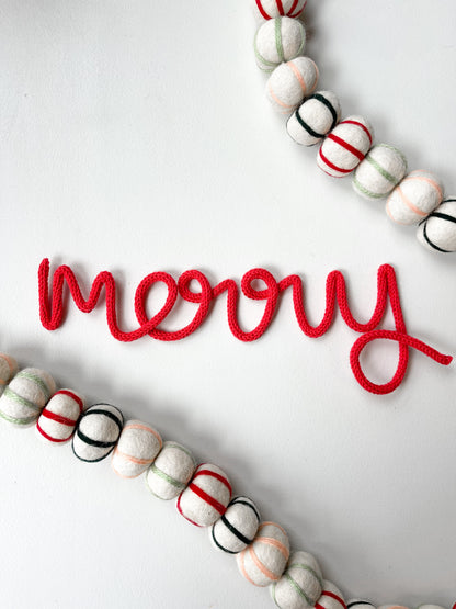 A red 'Merry' sign made of wire, with a white background, placed on a surface with Christmas decorations.
