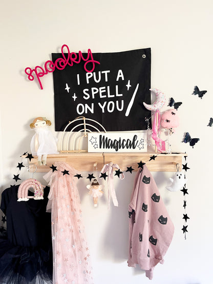 Black knitted wire ‘Spooky’ sign in neon pink angled over a black ‘I Put a Spell on You’ banner on a peg rail shelf in a little girl’s bedroom. Shelf is decorated with Halloween accents including a doll, wands, a black tutu, a cat sweatshirt, a pink glittery star cape, and a wire ‘Wands’ sign, creating a whimsical, festive, and magical Halloween display.