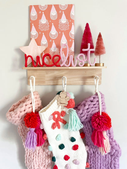 A custom wire 'nice list' sign with a red background, hanging on a wall. The sign has white text and decorative elements, and it's accompanied by a series of Christmas-themed knitted items in various colors.