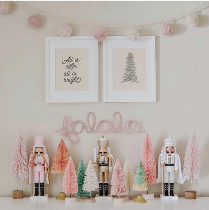 Blush pink ‘Fa La La’ knitted wire word in a Christmas setup with pink bottle brush trees, festive garland, nutcrackers and holiday art prints.