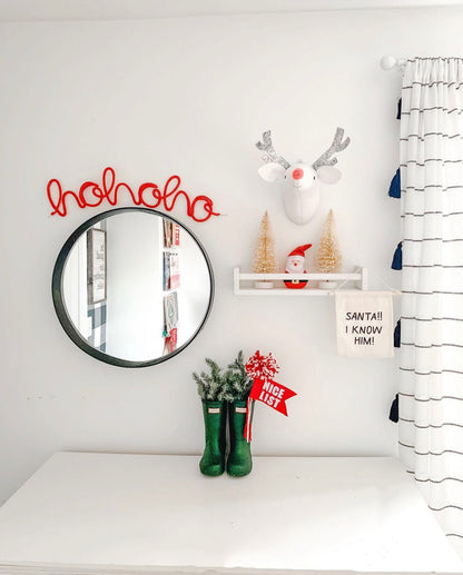 A decorative knitted wire art piece with 'hohoho' text and a reindeer design, placed on a white wall above a mirror, with Christmas-themed accessories around it.