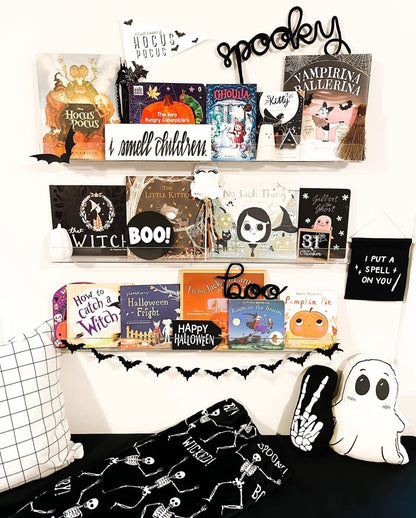 Children’s bedroom wall with three layered bookshelves filled with Halloween-themed books featuring pumpkins, witches, vampires, and ghosts. Black knitted wire word signs spelling ‘Spooky’ and ‘Boo’ are displayed on the shelves. Ghost garlands are draped across the shelves, and Halloween-themed accent pillows create a cozy reading spot with cushions below. Playful, festive, and whimsical seasonal décor for a child’s room or nursery.