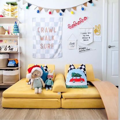A colorful kids room decorated for the holidays. A yellow play couch with festive pillows and stuffed animals. A garland, banners and knitted wire word in red 'hohoho' are on the wall'