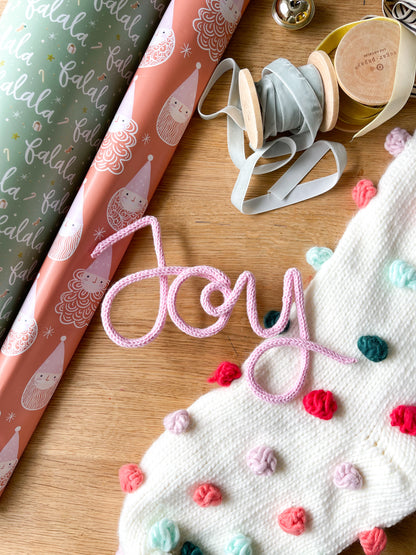 Handcrafted 'joy' sign with wire, fabric, and buttons on a wooden surface, featuring the word 'joy' embroidered in multicolor thread.