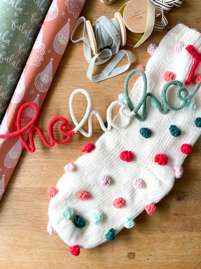 A knitted wire art decoration in the shape of the word 'hohoho' with a Christmas theme, featuring red, green, and white colors, placed on a wooden surface with holiday-themed wrapping paper and decorations.