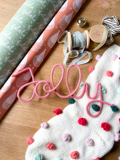 Flatlay of pink ‘Jolly’ knitted wire sign surrounded by Christmas stocking, wrapping ribbons, bells, and festive holiday decor.