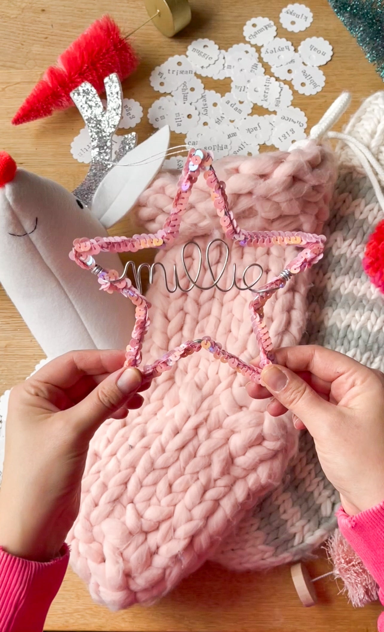 Two hands holding a pink sequin star ornament personalized with the name Millee in silver wire, with a pink Christmas stocking, whimsical Rudolph head mount, and other holiday decorations in the background.