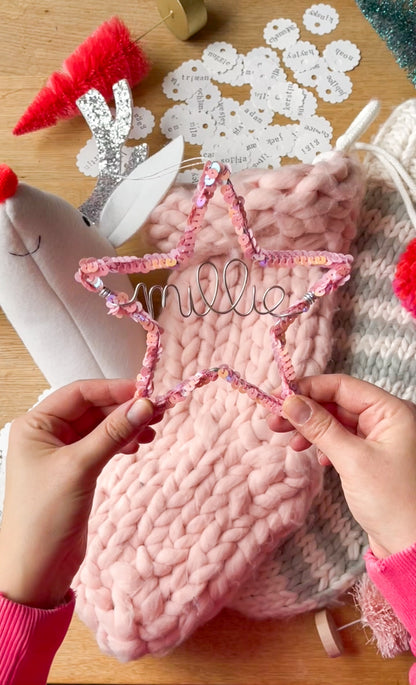Two hands holding a pink sequin star ornament personalized with the name Millee in silver wire, with a pink Christmas stocking, whimsical Rudolph head mount, and other holiday decorations in the background.