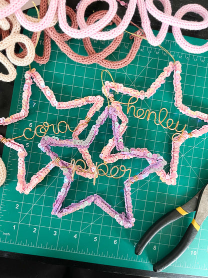 Three handmade sequin star ornaments on a green cutting mat — pink stars personalized for Cora and Henley, and a purple star for Piper — ready to be packaged as Christmas gifts, with bits of other wire names peeking into the frame.