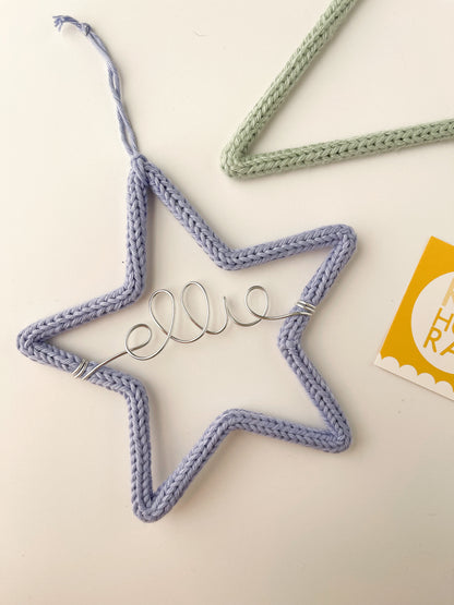 Star shaped ornament covered in periwinkle colored yarn with silver name 'ellie' in wire across the middle of it laying on white backdrop.