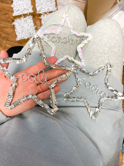 Hand holding a silver sequin star ornament personalized with the name Cali, with another silver star for Cairo and a white star for Caroline resting on a lap, surrounded by packaging materials, ready as handmade Christmas gifts.