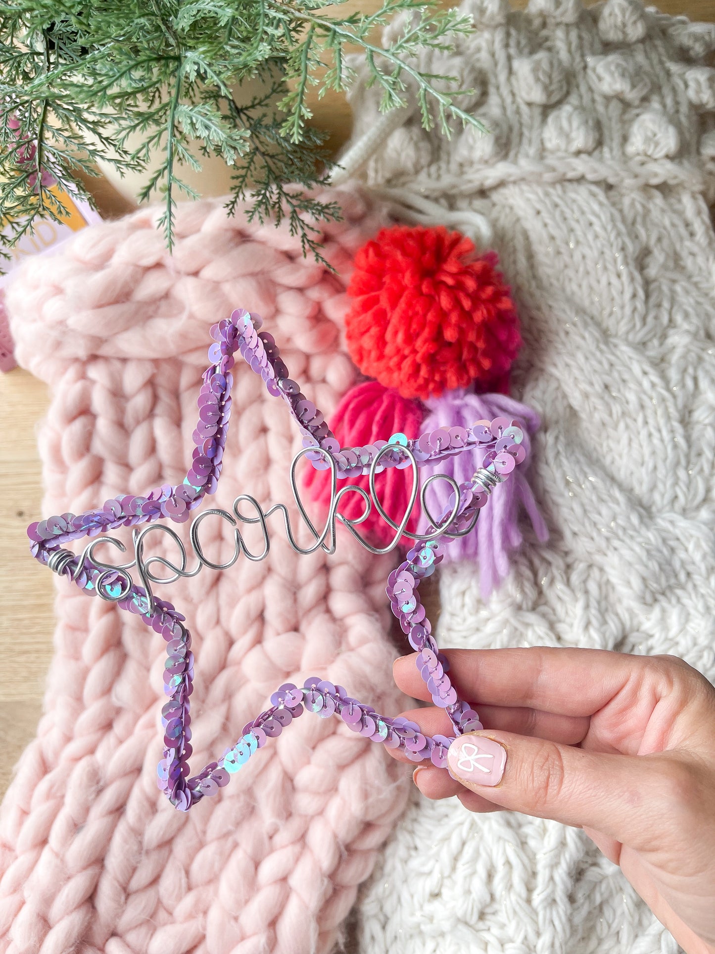 Hand holding a purple sequin star ornament with the word ‘Sparkle’ in silver wire, with a pink whimsical Christmas stocking, a white cozy chunky stocking, and a tree branch in the background, perfect for holiday decor.