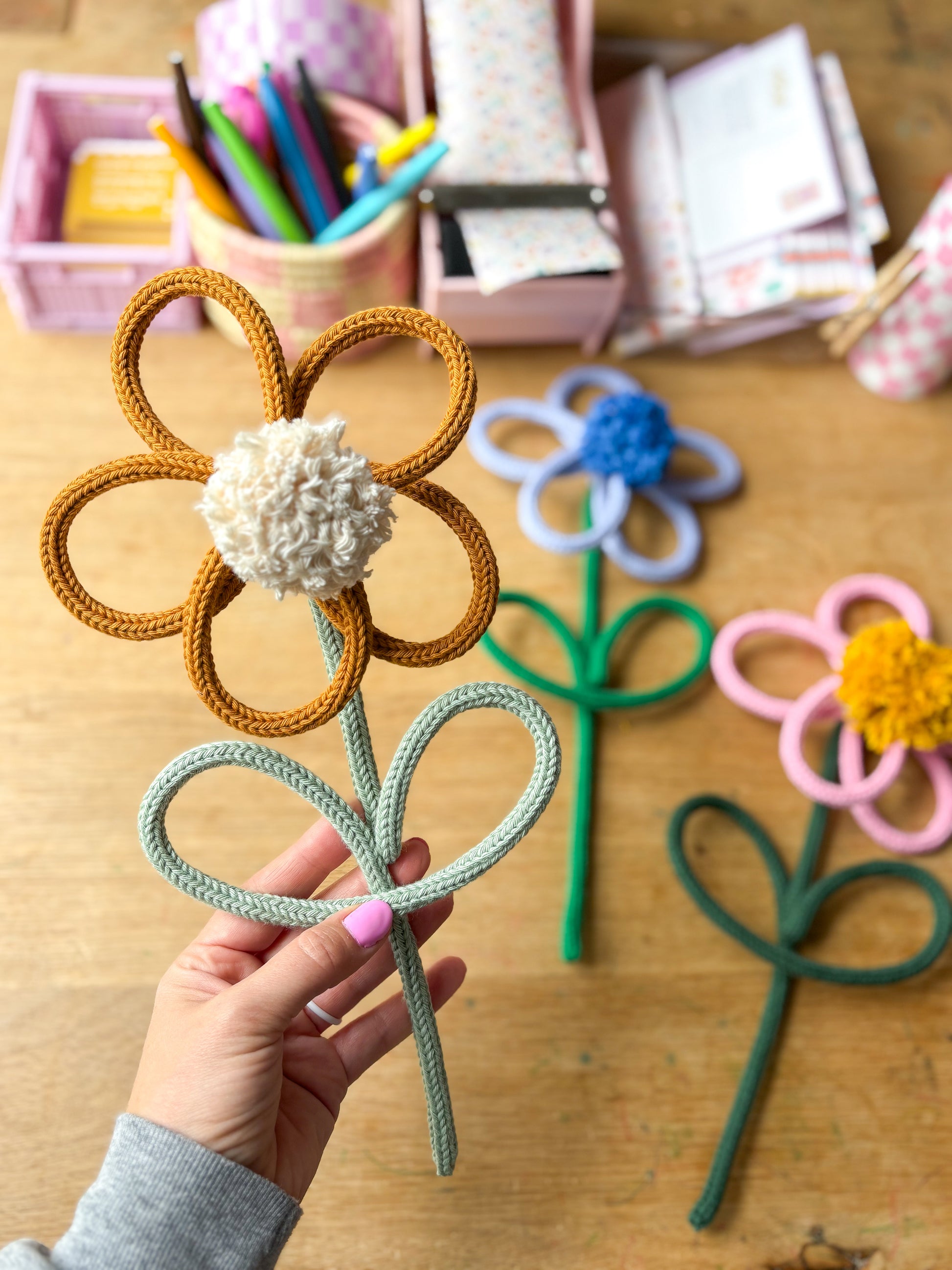 A hand holds a handmade rope flower \with mustard-brown petals, a white pom-pom center, and a sage green stem. Three more rope flowers in blue, yellow, and pink are arranged on a wooden table in the background, with craft supplies in pink bins.