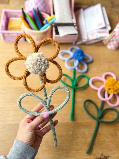 A hand holds a handmade rope flower \with mustard-brown petals, a white pom-pom center, and a sage green stem. Three more rope flowers in blue, yellow, and pink are arranged on a wooden table in the background, with craft supplies in pink bins.